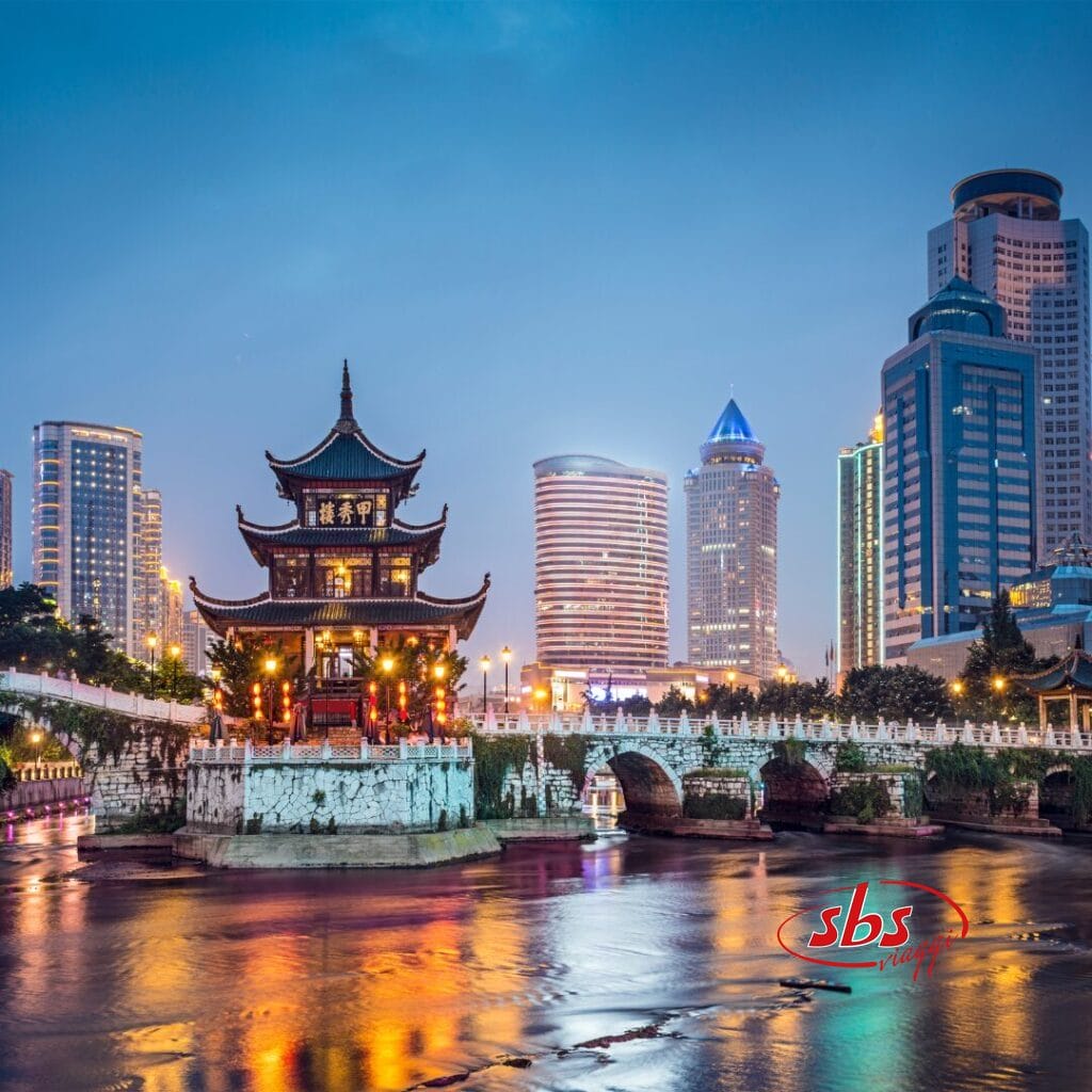 Un tradizionale padiglione cinese accanto a un ponte di pietra illuminato, circondato da moderni grattacieli, con le luci della città che si riflettono sul fiume al tramonto: una scena perfetta di Cina Classica, ideale per la tua avventura nel Tour Balla coi Panda.