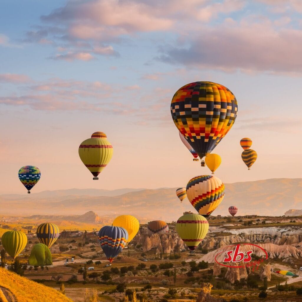 Diverse mongolfiere colorate fluttuano sopra un paesaggio roccioso all'alba, con montagne in lontananza e vegetazione sparsa visibili in basso: una vista mozzafiato che ricorda un'indimenticabile avventura del Tour Istanbulissima.