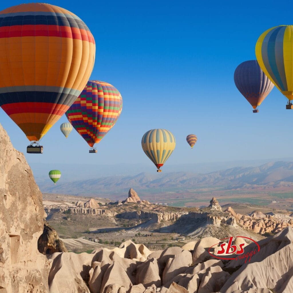 Numerose mongolfiere colorate sorvolano il paesaggio roccioso della Cappadocia, con le montagne sullo sfondo e un cielo azzurro e limpido: uno scenario mozzafiato che puoi ammirare durante un'avventura con il Tour Cappadocia.