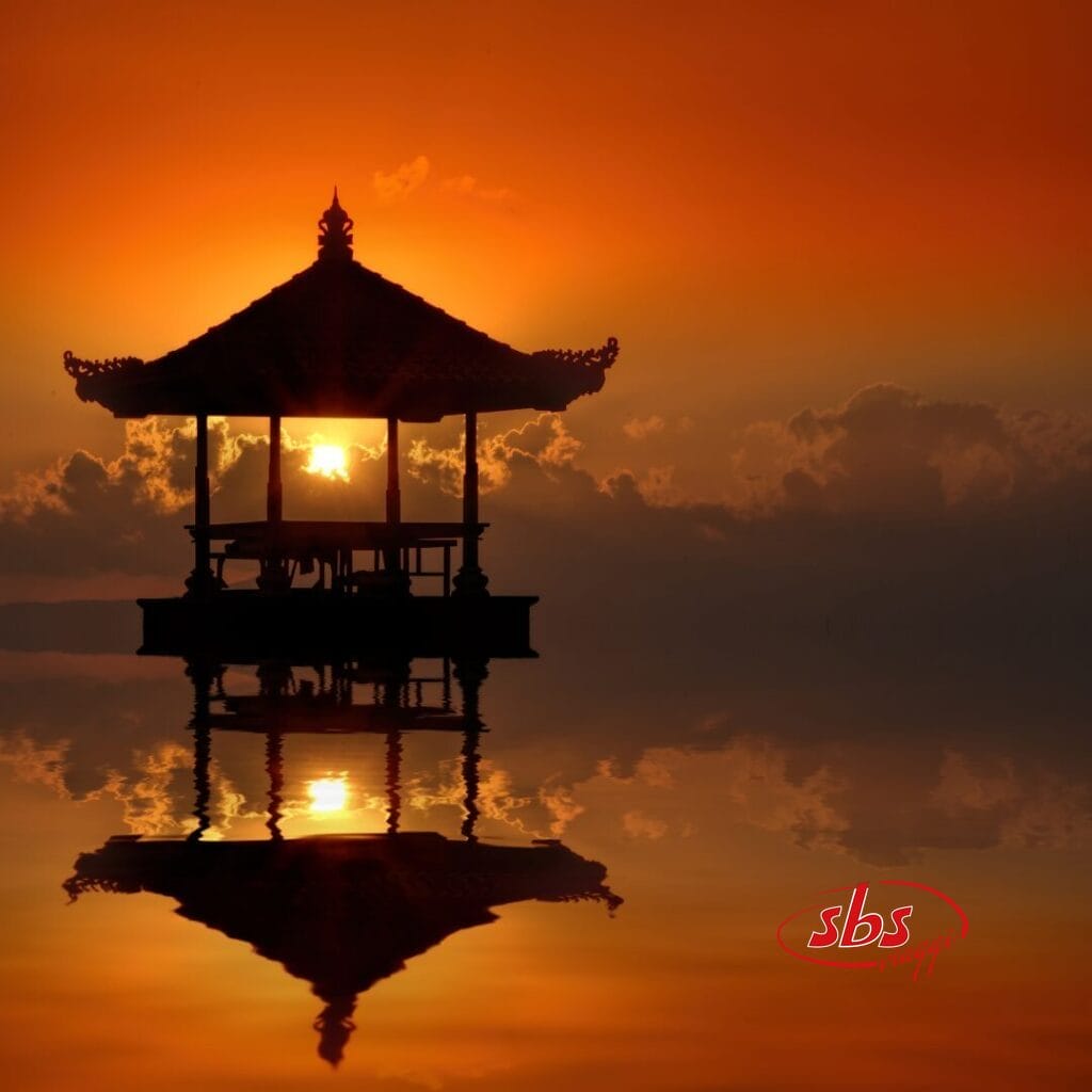La sagoma di un tradizionale padiglione balinese si riflette nell'acqua calma al tramonto, con un cielo arancione e nuvole sullo sfondo: perfetto per il tuo prossimo tour su Instagram.