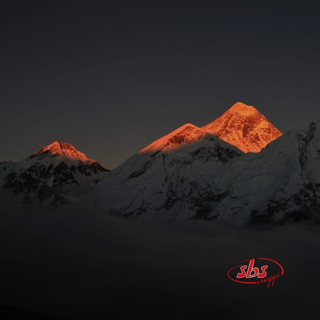 Le cime innevate delle montagne, illuminate dai raggi arancioni dei raggi del sole, si ergono sopra le nuvole nella Valle degli Dèi, contro un cielo scuro: un momento indimenticabile di ogni avventura del Tour Nepal.