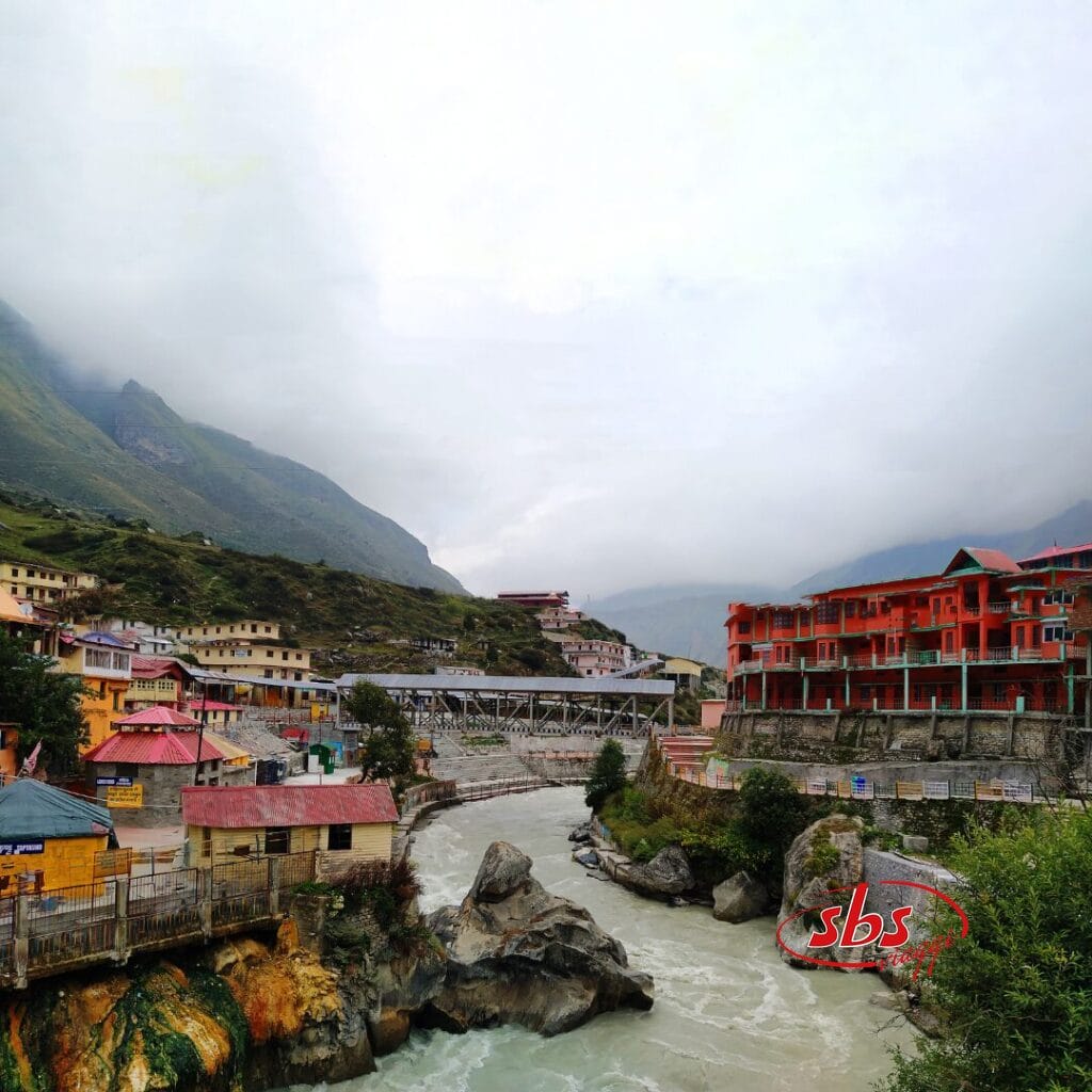 Un fiume scorre attraverso una cittadina di montagna in Nepal con edifici colorati e un ponte di metallo, circondata da colline nebbiose e cieli nuvolosi, catturando il fascino sereno spesso visibile durante il Festival di Dashain.