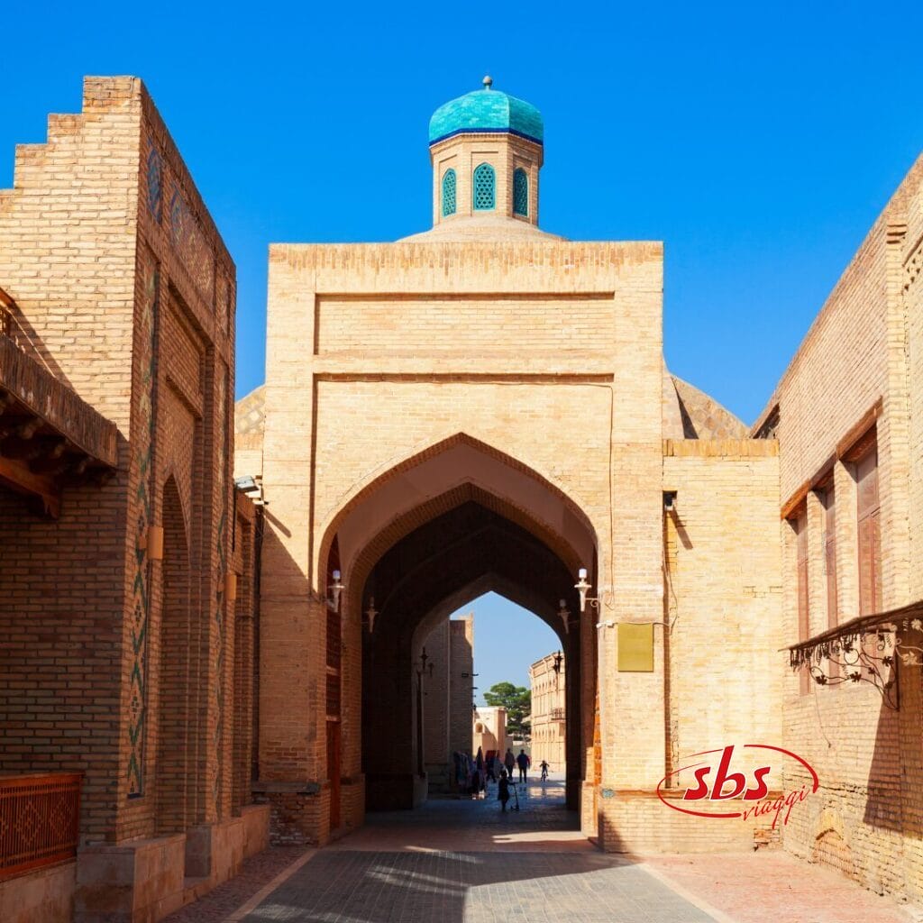 Uno storico arco in mattoni con una cupola blu in cima, fiancheggiato da edifici, sotto un cielo azzurro e limpido, con persone che camminano lungo il passaggio: uno spettacolo iconico di qualsiasi tour in Uzbekistan.