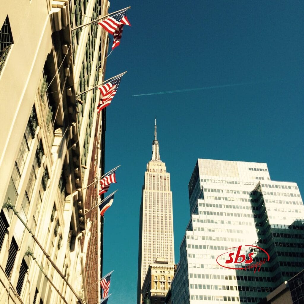 Numerose bandiere americane sono appese a un edificio con l'Empire State Building e altri grattacieli sullo sfondo, catturando la vivace atmosfera di Ferragosto a New York sotto un limpido cielo azzurro.