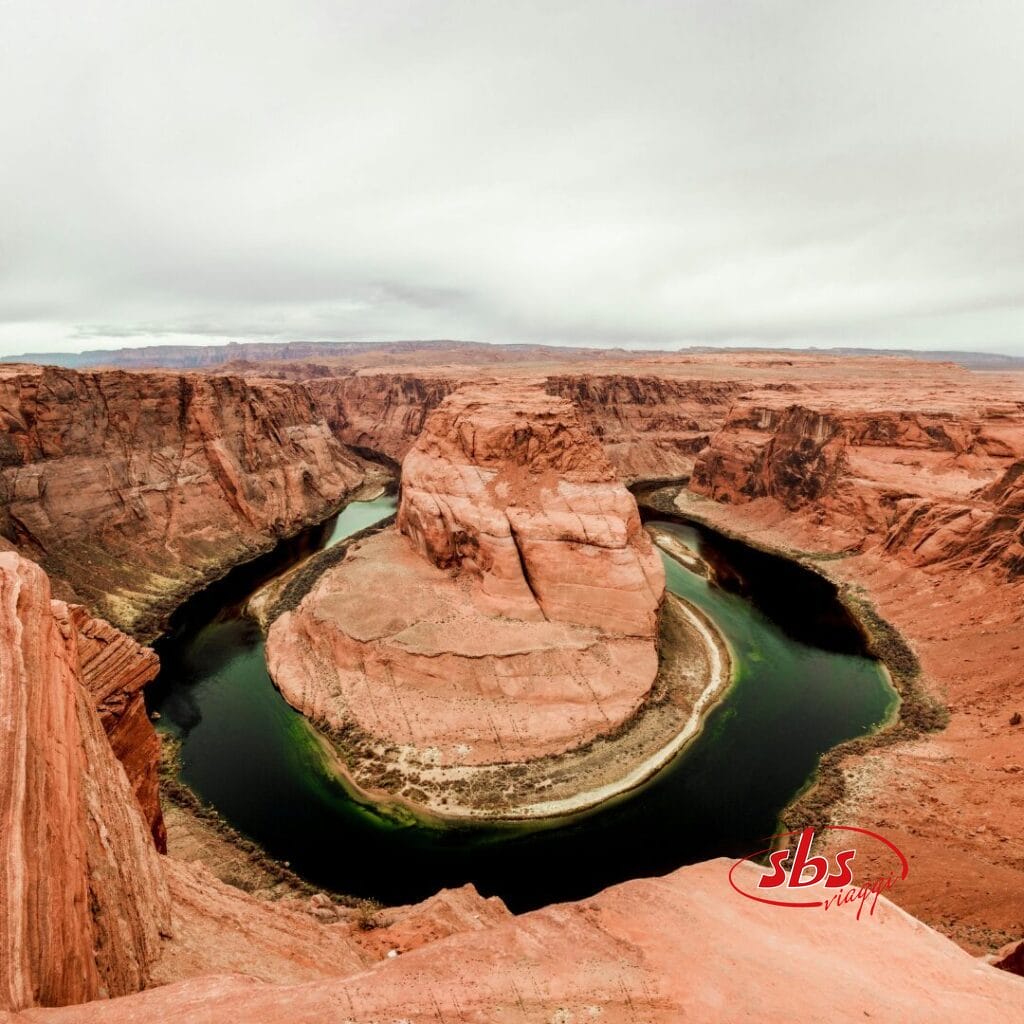 Un'ampia vista di Horseshoe Bend, dove il fiume Colorado si curva attorno a una formazione rocciosa di arenaria sotto un cielo nuvoloso in un paesaggio desertico, fondendo la bellezza naturale con la modernità e le tradizioni delle iconiche capitali del turismo.