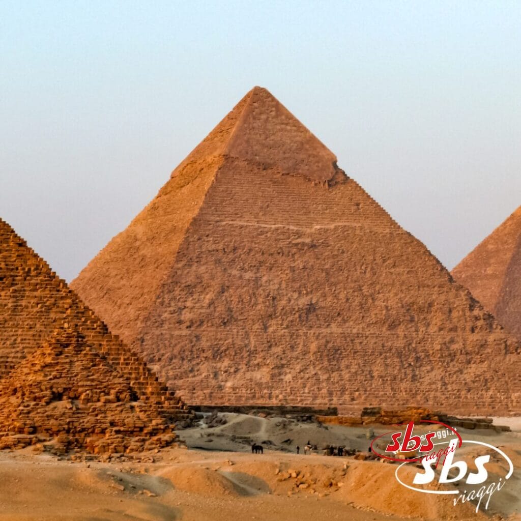 Tre grandi piramidi si ergono nel deserto al tramonto, vicino al Cairo, con un gruppo di persone e cammelli nelle vicinanze.