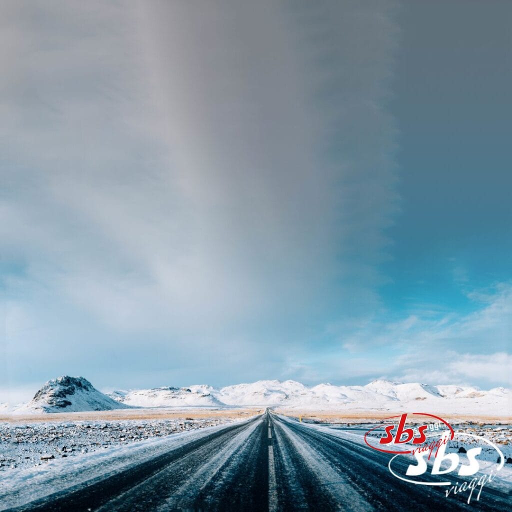 Una strada dritta conduce alle montagne innevate dell'Islanda sotto un cielo azzurro con nuvole; nell'angolo compaiono la scritta