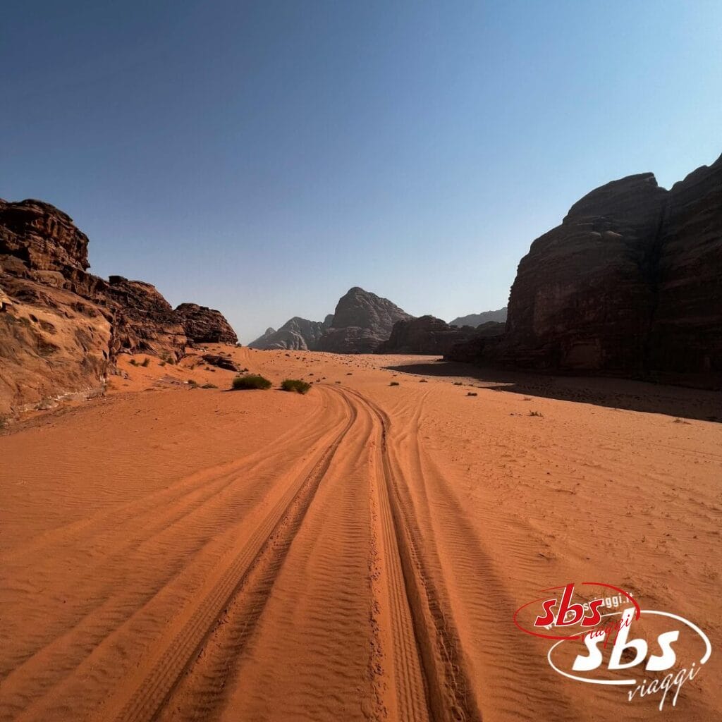 Tracce di pneumatici nella sabbia rossa del deserto con scogliere rocciose sotto un cielo azzurro; Nell'angolo il logo sbs viaggi, che evoca il Gran Tour della Giordania per un viaggio completo.