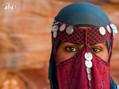 Una persona indossa abiti tradizionali e un velo decorato con monete, che evocano il patrimonio dell'Arabia Saudita, in piedi su uno sfondo sfocato.