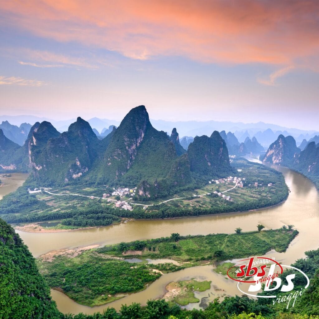 Un fiume serpeggia tra le lussureggianti colline verdi e le montagne carsiche del Guizhou sotto un cielo colorato al tramonto, con piccoli villaggi lungo la riva: una scena classica di Cina d’Autore.