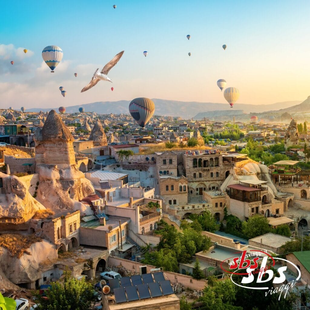 Una vista panoramica delle formazioni rocciose e degli edifici della Cappadocia con mongolfiere, un gabbiano nel cielo e il logo di SBS Viaggi nell'angolo: perfetta per il tuo prossimo tour di Istanbulissima o della Cappadocia.
