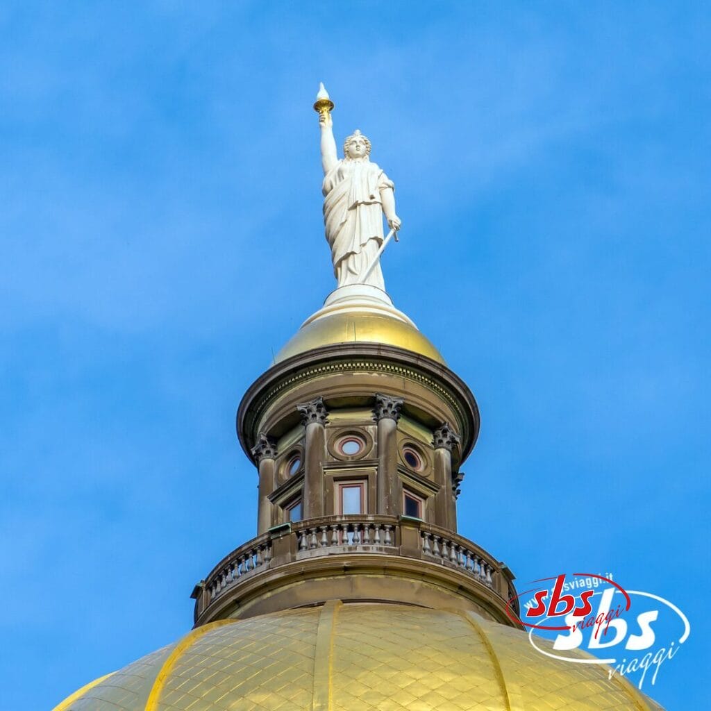 Primo piano di un edificio con cupola dorata in Georgia, sormontato da una statua bianca che regge una torcia, sullo sfondo di un cielo azzurro e terso.