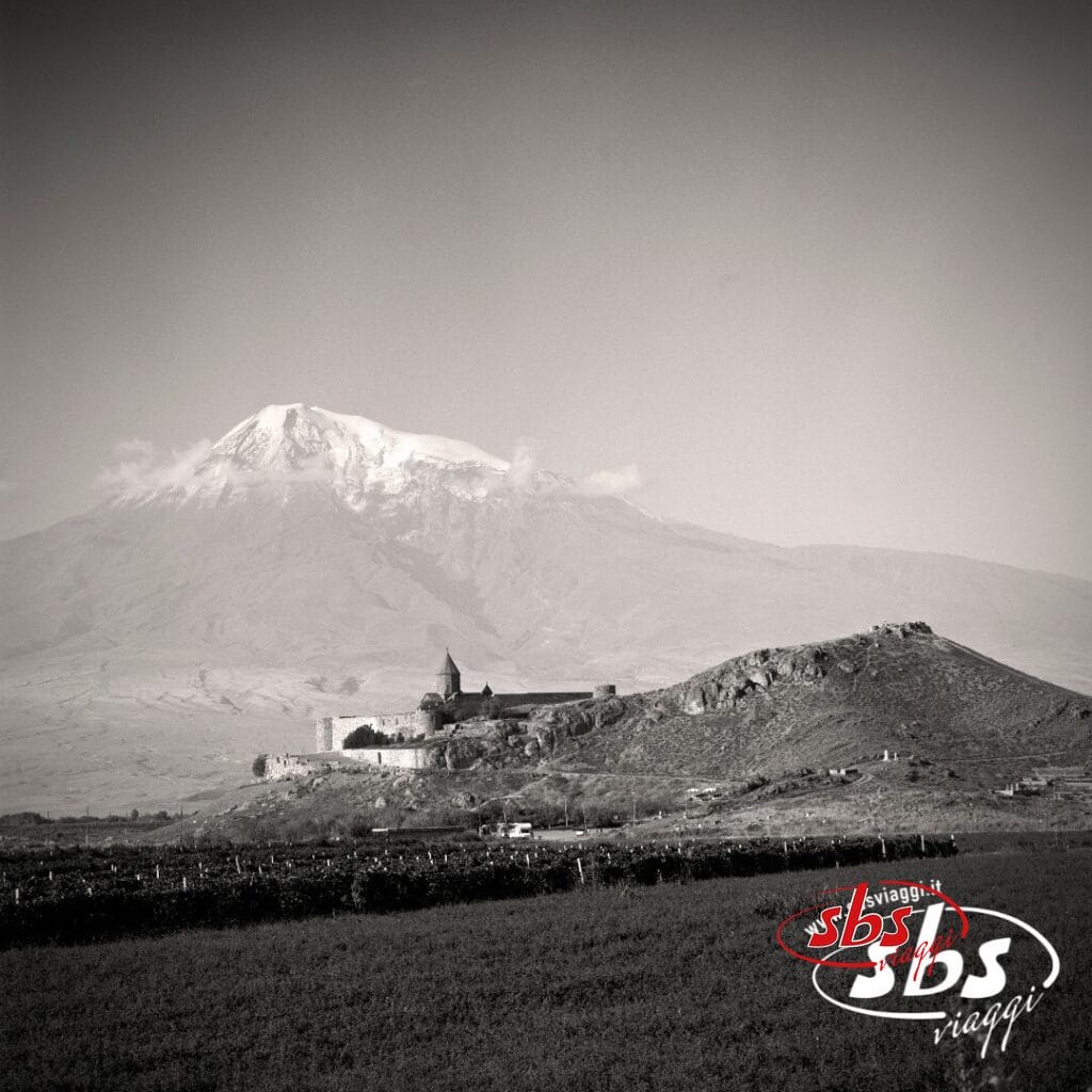 Una foto in bianco e nero mostra un monastero su una collina in Georgia, con una montagna innevata sullo sfondo. Il logo di