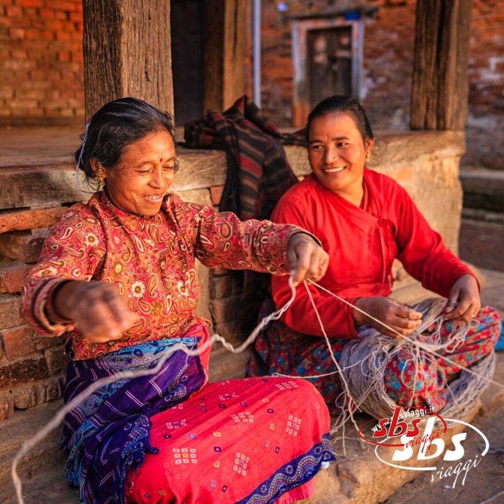 Due donne siedono su un portico rustico nella Valle degli Dei, sorridenti mentre lavorano con il filato. Una dipana i fili mentre l'altra osserva, godendosi un momento di pace all'aria aperta. Visitate il Nepal e scoprite scenari così sereni.