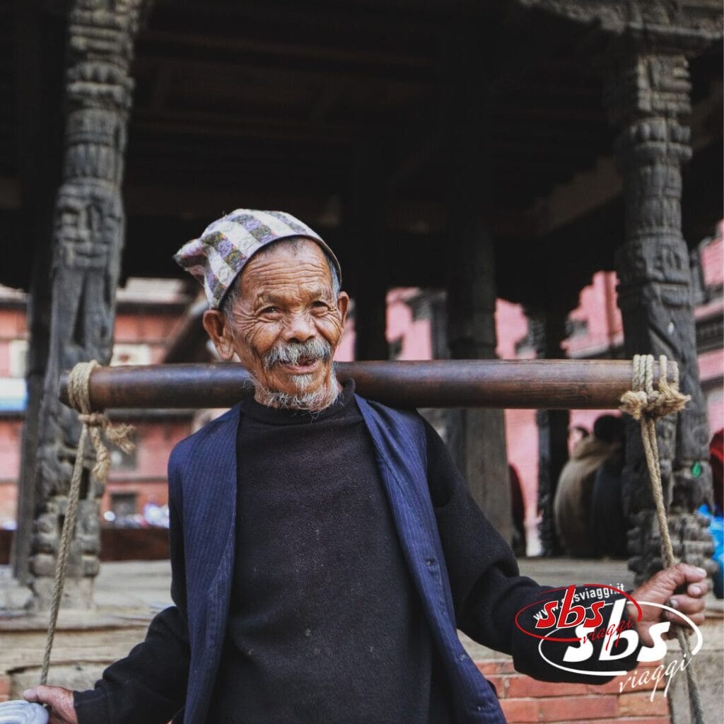Un uomo anziano che indossa un cappello e un gilet decorati porta un palo di legno sulle spalle in un cortile nepalese con pilastri in pietra scolpita, evocando la ricca cultura ammirata durante un tour del Triangolo d'Oro.