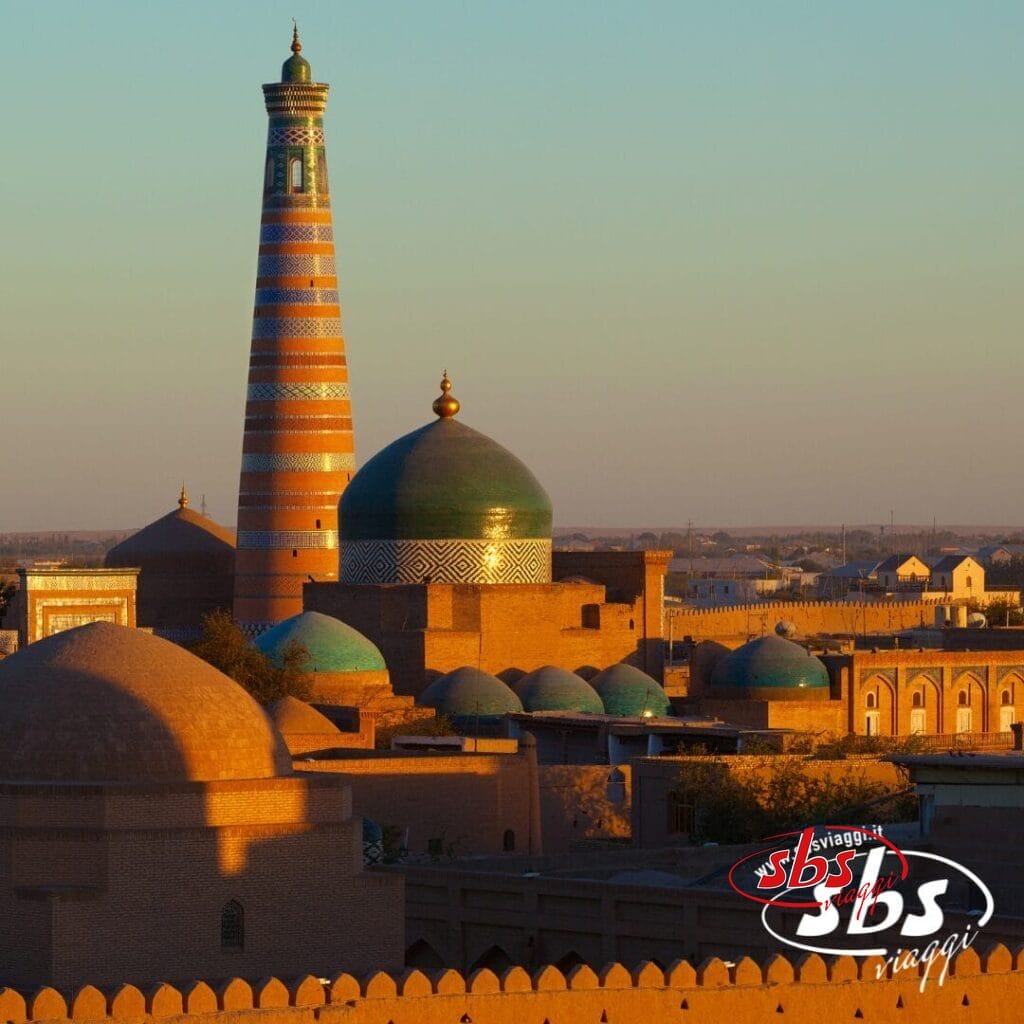 Vista soleggiata di edifici storici con cupole turchesi e un alto minareto a strisce a Khiva, in Uzbekistan, perfetta per il tuo prossimo tour in Uzbekistan con il logo SBS Viaggi nell'angolo.