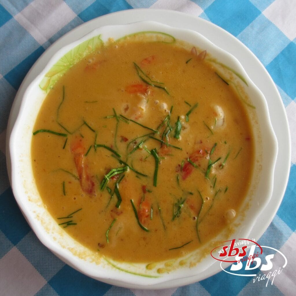Una ciotola di cremosa zuppa all'arancia con erbe aromatiche e pezzi di pomodoro a vista, adagiata su un piatto bianco sopra una tovaglia a quadri bianchi e blu, evoca i sapori confortanti che spesso si possono scoprire durante un tour Grantour o Cambogia.