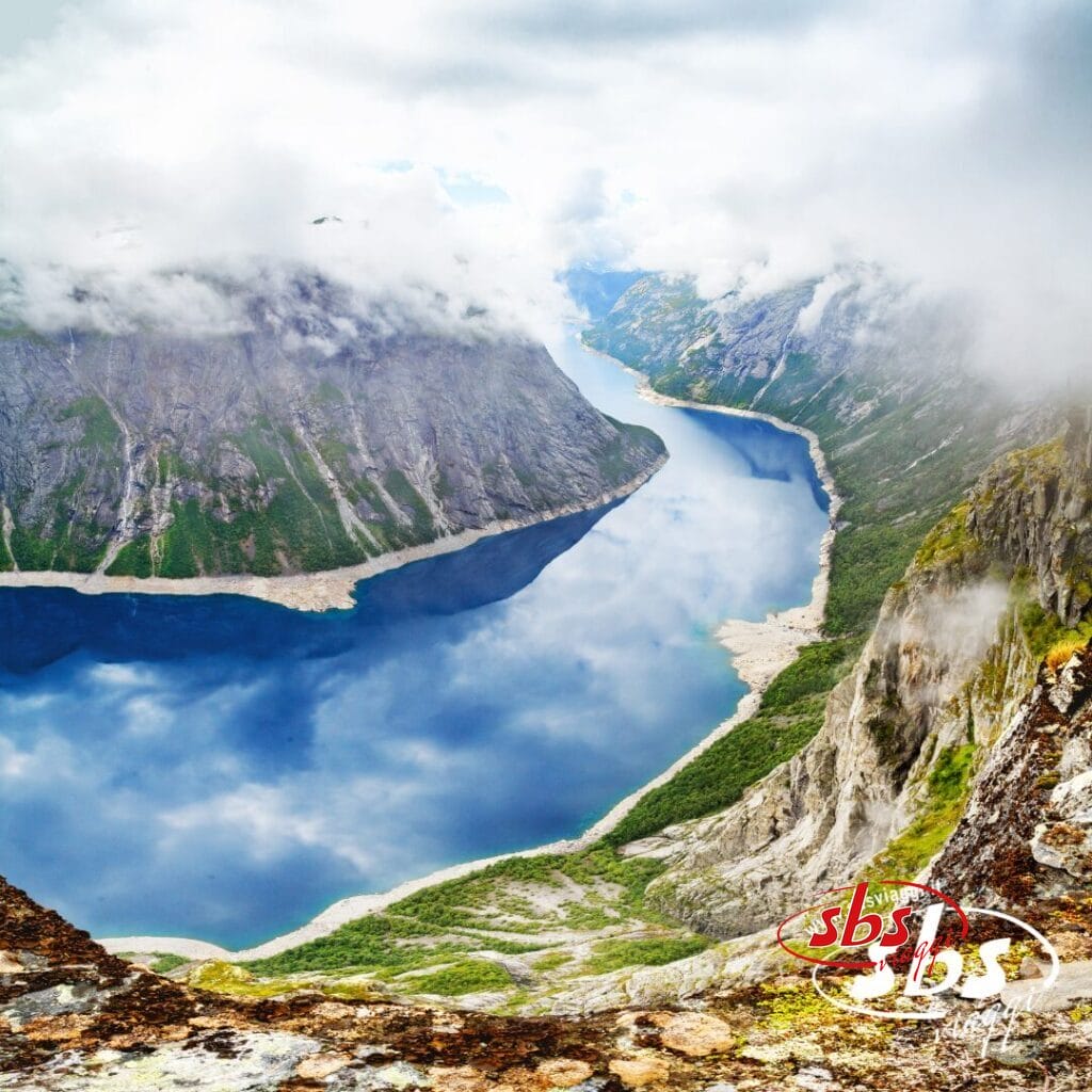 Un fiume azzurro e limpido serpeggia tra montagne ripide e rocciose ricoperte di verde, con nuvole che ne nascondono parzialmente le cime: uno scenario perfetto per un Tour Fiordi Meravigliosi con accompagnatore dall’Italia.