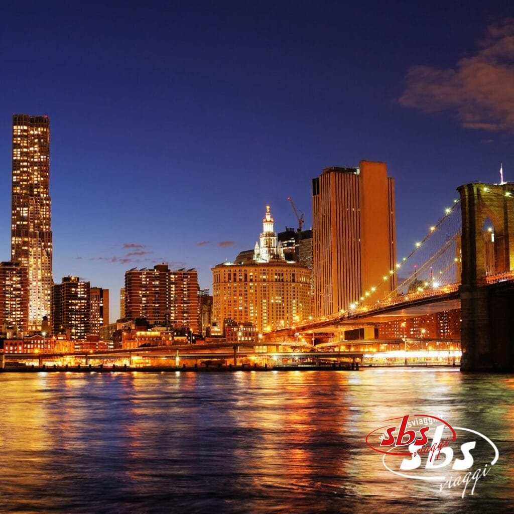 Vista notturna del ponte di Brooklyn e dello skyline del centro di Manhattan con gli edifici illuminati che si riflettono sull'acqua, perfetta per vivere Ferragosto a New York con il tuo accompagnatore dall'Italia.