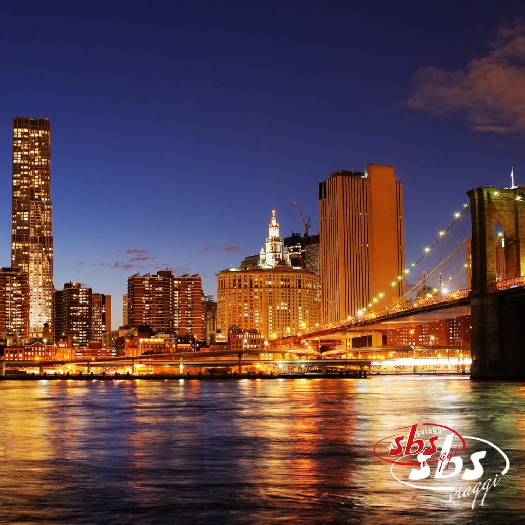 Uno scatto urbano notturno con edifici illuminati e il ponte di Brooklyn riflesso nell'acqua sotto un cielo blu scuro, che cattura la magia di Settembre a New York.