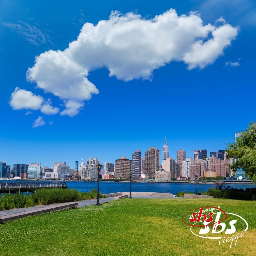 Un parco con erba verde si affaccia su un fiume e sullo skyline di New York City sotto un cielo azzurro con nuvole sparse; nell'angolo compaiono il logo 