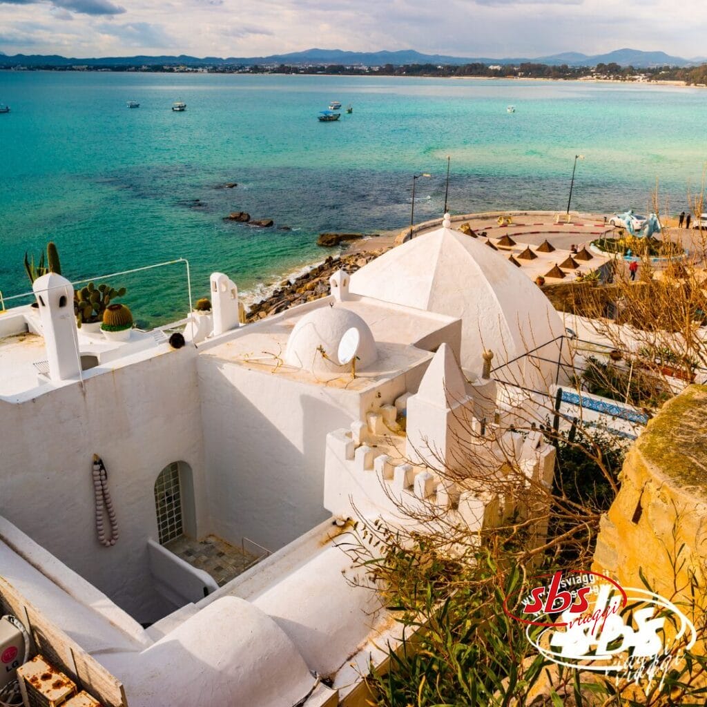 Un edificio bianco in stile mediterraneo che si affaccia sul mare turchese e sulla spiaggia, con barche e ombrelloni sotto un cielo nuvoloso: ammirate questa vista durante il Tour Autentica Tunisia.