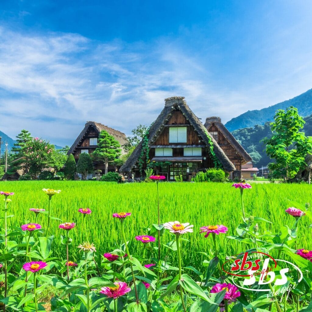 Le tradizionali case con il tetto di paglia in Giappone sorgono dietro una risaia verde, con fiori colorati in primo piano e montagne sullo sfondo sotto un cielo azzurro.