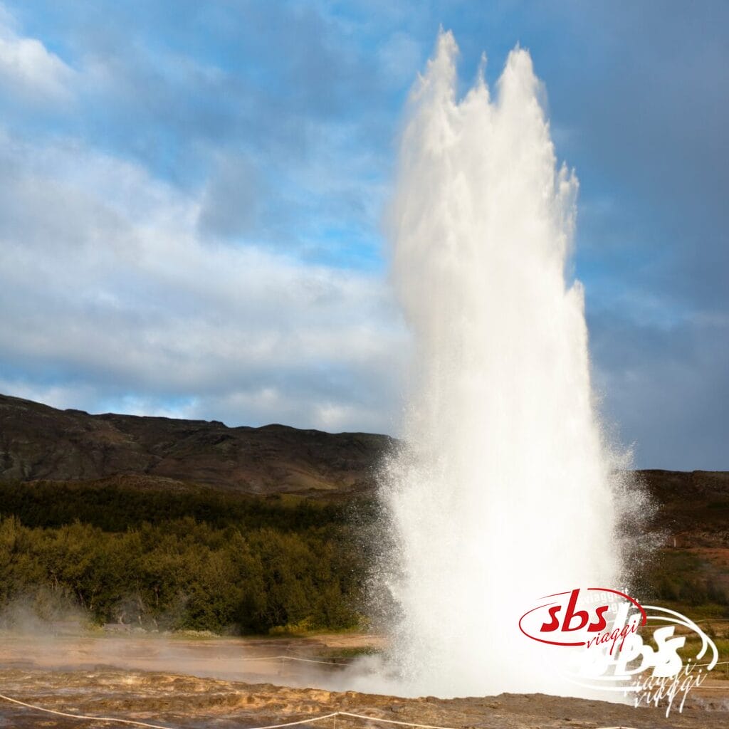 Un potente geyser erutta, sparando acqua in aria contro un cielo nuvoloso e colline in lontananza: uno spettacolo maestoso sui Meravigliosi Altopiani.