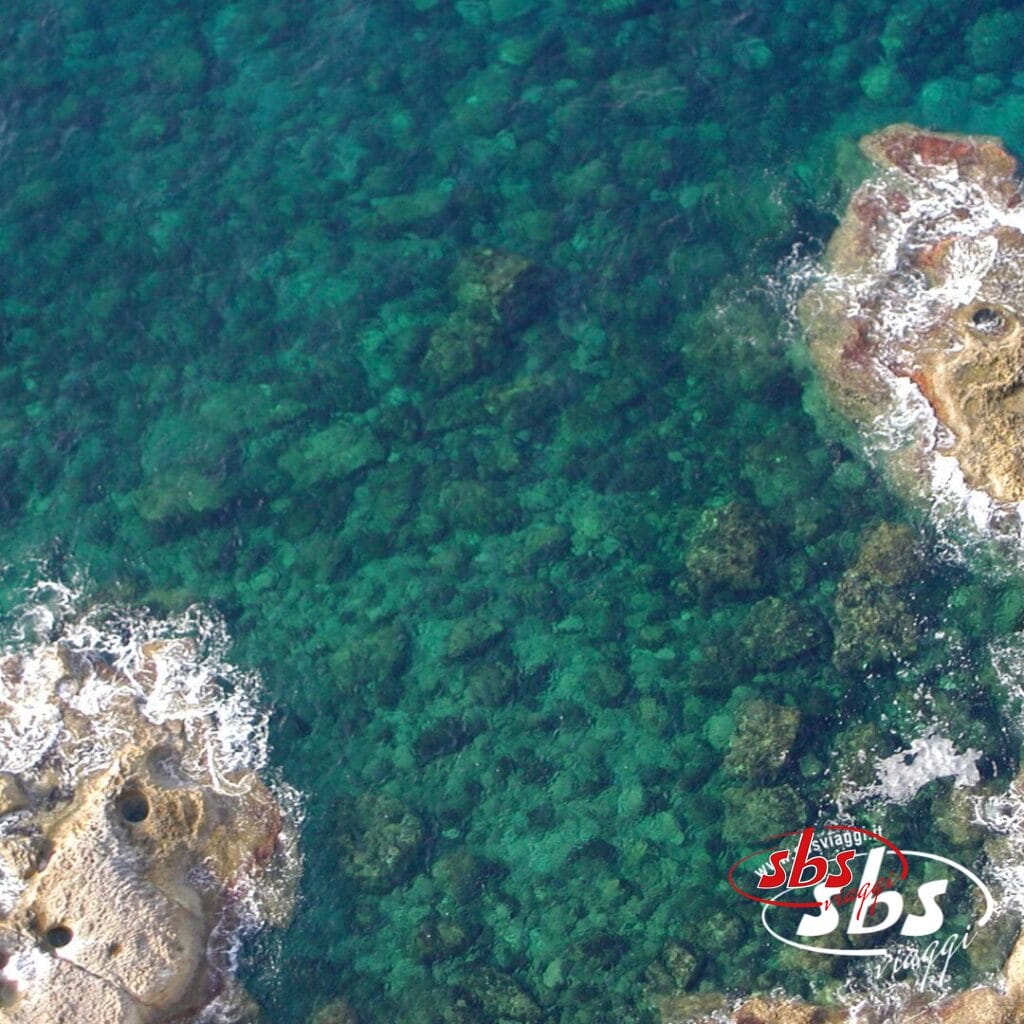 Vista aerea di acque turchesi e cristalline con formazioni rocciose e onde bianche, logo SBS Viaggi visibile: scopri la bellezza di Case Vacanza Lampedusa.