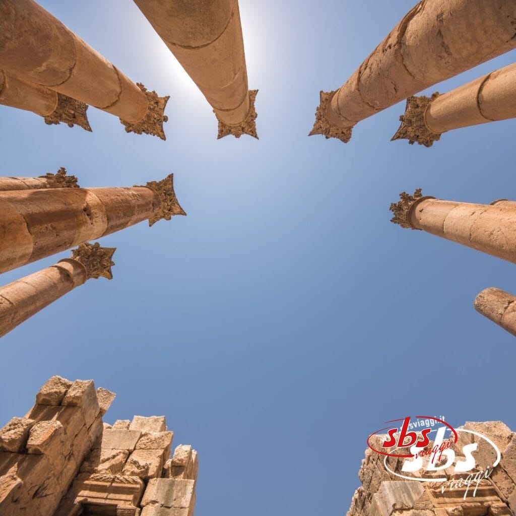 Antiche colonne di pietra si ergono verso il cielo azzurro e terso della Giordania, viste dal basso con la luce del sole alle spalle: uno spettacolo mozzafiato durante un Gran Tour.
