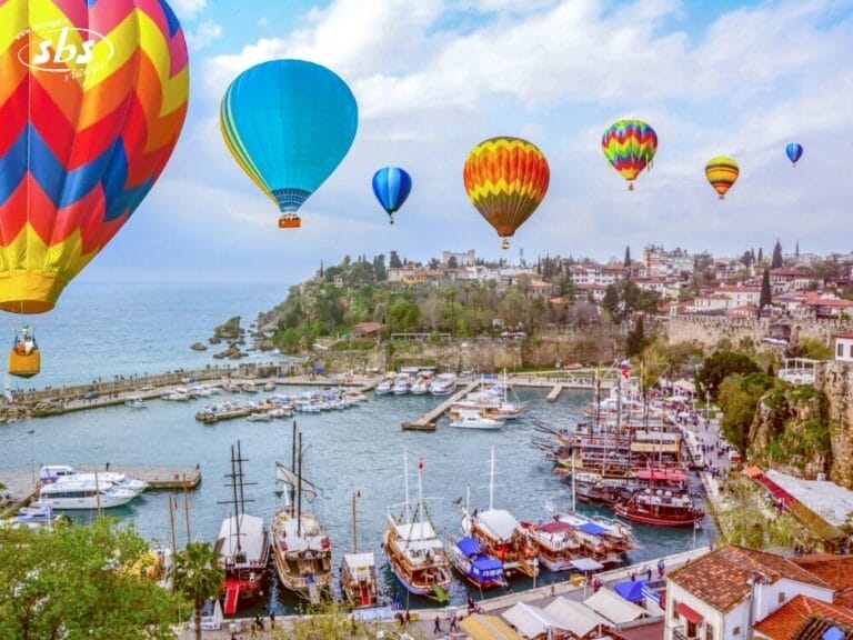 Colorate mongolfiere fluttuano sopra una città costiera con barche ormeggiate e un porto vivace, evocando lo spirito del Tour Sapori del Mediterraneo sotto un cielo parzialmente nuvoloso.