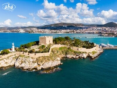 Vista aerea del castello in pietra e del faro su una piccola isola di Mare, con la Torre Kervansaray visibile in lontananza tra le acque blu, il paesaggio urbano e le colline sotto un cielo parzialmente nuvoloso.