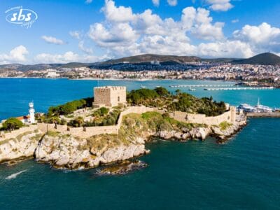 Vista aerea del castello in pietra e del faro su una piccola isola di Mare, con la Torre Kervansaray visibile in lontananza tra le acque blu, il paesaggio urbano e le colline sotto un cielo parzialmente nuvoloso.