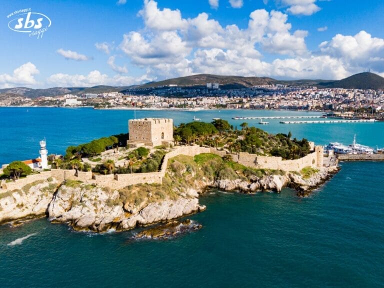 Vista aerea del castello in pietra e del faro su una piccola isola di Mare, con la Torre Kervansaray visibile in lontananza tra le acque blu, il paesaggio urbano e le colline sotto un cielo parzialmente nuvoloso.