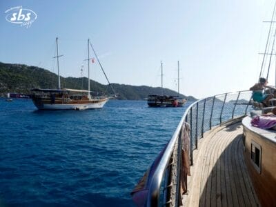 Tre barche a vela sono ancorate su un mare calmo e blu, vicino a una costa collinare e verde, con parte del ponte e della ringhiera di un caicco visibili in primo piano, forse in attesa della prossima avventura del Tour Kervansaray.
