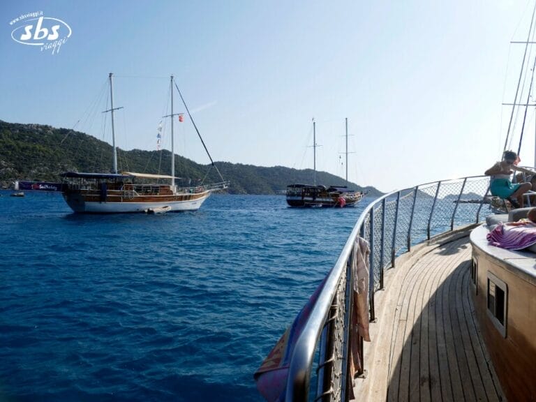 Tre barche a vela sono ancorate su un mare calmo e blu, vicino a una costa collinare e verde, con parte del ponte e della ringhiera di un caicco visibili in primo piano, forse in attesa della prossima avventura del Tour Kervansaray.