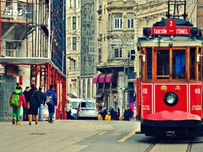 Un tram rosso d'epoca con la scritta "Taksim-Tünel" percorre una strada cittadina fiancheggiata da edifici storici e pedoni, offrendo un'esperienza unica del tour Istanbulissima.