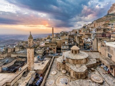 Edifici storici in pietra con tetti a cupola e minareti in una città vecchia al tramonto, sotto un cielo nuvoloso spettacolare: scopri i Tesori della Mesopotamia durante il tuo prossimo tour, con montagne e terreni aperti che si estendono sullo sfondo.