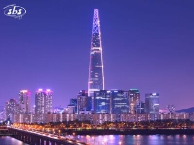 Vista notturna dello skyline di una città con un alto grattacielo illuminato circondato da altri edifici, con un ponte e un fiume in primo piano: una tappa obbligata del tuo Tour Corea del Sud.