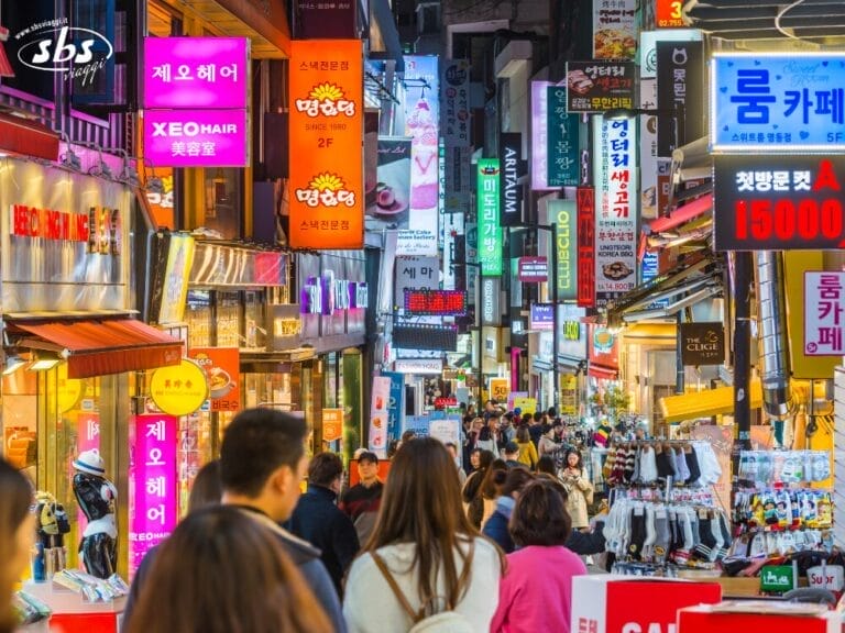 Una strada trafficata della Corea del Sud piena di gente e di insegne luminose in coreano che pubblicizzano negozi, ristoranti e saloni di bellezza: uno scenario vivace, perfetto per qualsiasi avventura del Gran Tour Corea.
