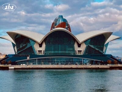 Moderno edificio architettonico con grandi tetti bianchi a forma di petalo, situato accanto a uno specchio d'acqua sotto un cielo nuvoloso, che mette in mostra influenze di design provenienti dall'Azerbaijan e dalla Georgia.