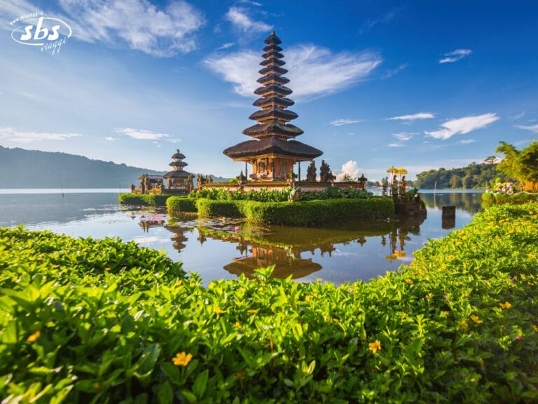 Un tradizionale tempio balinese con tetti a più livelli sorge su una piccola isola circondata da acqua, vegetazione lussureggiante e montagne sotto un cielo terso e luminoso: uno scenario perfetto per Instagram da immortalare durante il tuo tour dell'isola.
