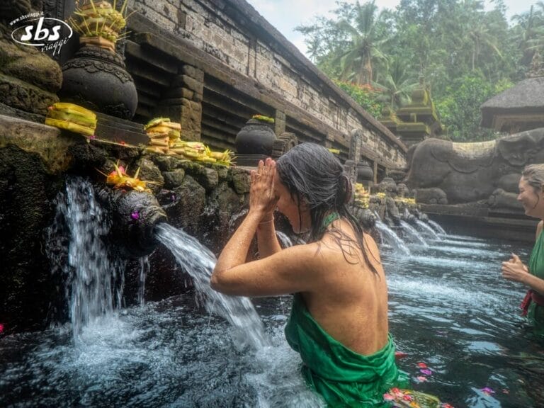 Una donna è in piedi in una piscina di pietra in un tempio di Bali, con le mani giunte in preghiera sotto un getto d'acqua, con offerte sopra i getti e un'altra persona nelle vicinanze: una scena serena, perfetta per qualsiasi viaggio.