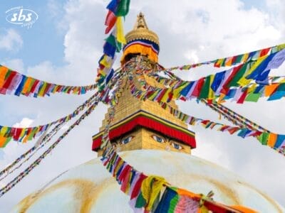 Un grande stupa con occhi di Buddha e colorate bandiere di preghiera che sventolano dalla sua torre contro un cielo parzialmente nuvoloso, invitandovi a visitare il Nepal e a scoprire le meraviglie mistiche di questa Valle degli Dèi.