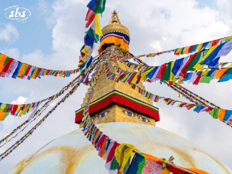 Un grande stupa con occhi di Buddha e colorate bandiere di preghiera che sventolano dalla sua torre contro un cielo parzialmente nuvoloso, invitandovi a visitare il Nepal e a scoprire le meraviglie mistiche di questa Valle degli Dèi.
