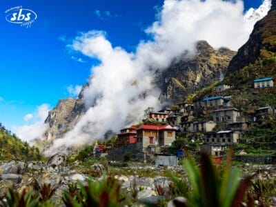 Un villaggio himalayano in Nepal, con case colorate, sorge ai piedi di montagne scoscese e nebbiose, sotto un cielo parzialmente nuvoloso. Una vegetazione lussureggiante riempie il primo piano, catturando il fascino di questa Valle degli Dei, soprattutto durante il Festival di Dashain.