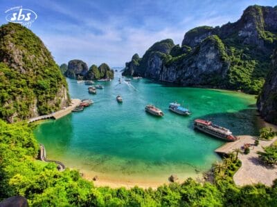 Una baia pittoresca con acqua color smeraldo, numerose imbarcazioni turistiche del Gran Tour del Vietnam, scogliere calcaree verdi e una piccola spiaggia sabbiosa circondata da una vegetazione lussureggiante.