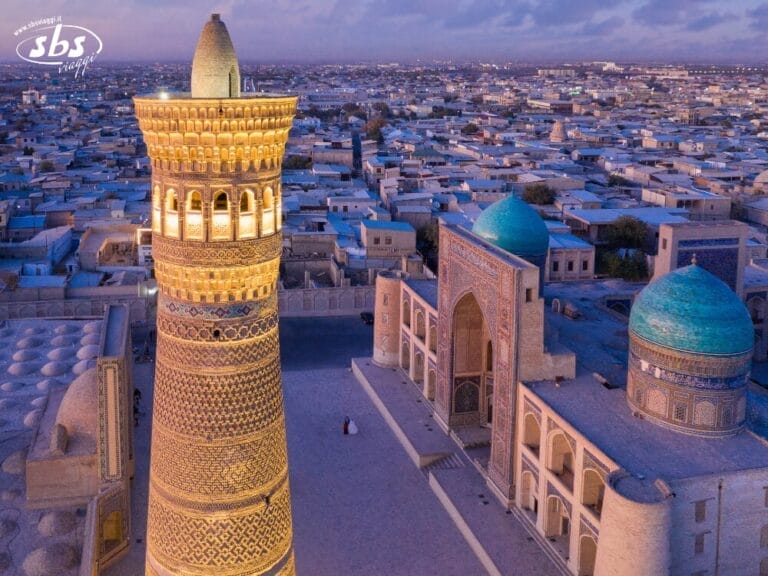 Vista aerea del minareto Kalyan illuminato e della moschea con cupole blu a Bukhara, in Uzbekistan, circondata da edifici densamente popolati al tramonto: una destinazione affascinante del Grantour.