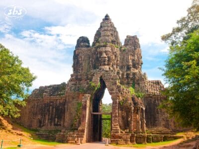Antica porta in pietra con grandi volti scolpiti ad Angkor Thom in Cambogia, circondata da alberi e sotto un cielo parzialmente nuvoloso: una tappa obbligata di qualsiasi tour Grantour o in Cambogia.