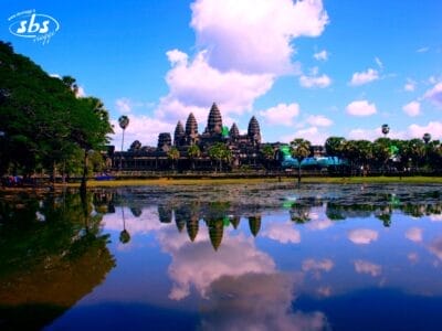 Il tempio di Angkor Wat si riflette in uno stagno immobile sotto un cielo azzurro brillante con nuvole sparse, circondato da alberi verdi e palme: uno dei momenti salienti di ogni esperienza del Tour Cambogia.